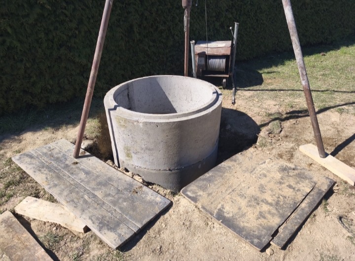 Betonová skruž osazená v zemi pod trojnožkou s vrátkem, vedle odložené dřevěné kryty