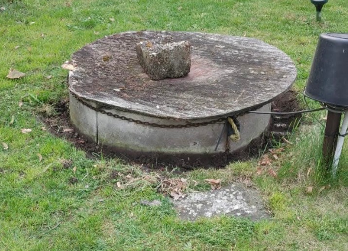 Betonový poklop studny na trávníku, částečně zarostlý mechem, s kamenem jako přítěží
